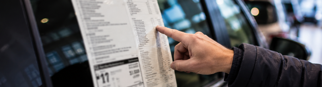 Person in a dark jacket points at a price sheet stuck to a car window in a showroom, inspecting the vehicle's details.