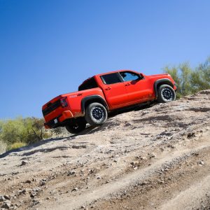 Red 2026 Nissan Frontier PRO-4X crew cab pickup truck descending a rocky desert trail under a clear blue sky.
