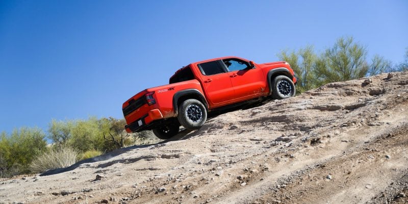 Red 2026 Nissan Frontier PRO-4X crew cab pickup truck descending a rocky desert trail under a clear blue sky.