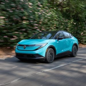 Turquoise 2026 Nissan Leaf driving down a tree-lined road, showing the redesigned crossover-coupe body and closed front fascia.