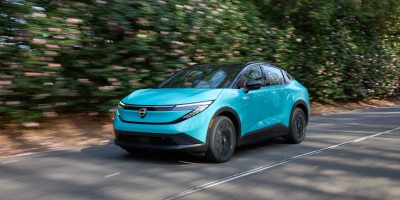 Turquoise 2026 Nissan Leaf driving down a tree-lined road, showing the redesigned crossover-coupe body and closed front fascia.