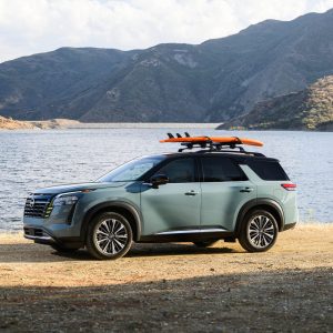 Sage green 2026 Nissan Pathfinder Platinum parked on a rocky lakeshore with a mountain backdrop and an orange surfboard mounted on the roof rack.