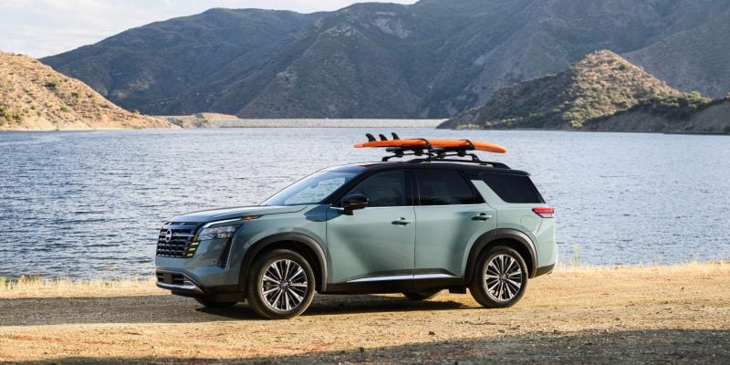 Sage green 2026 Nissan Pathfinder Platinum parked on a rocky lakeshore with a mountain backdrop and an orange surfboard mounted on the roof rack.
