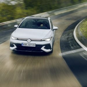 White 2026 Volkswagen Golf GTI with red grille accent driving through a corner on a race track under an overcast sky