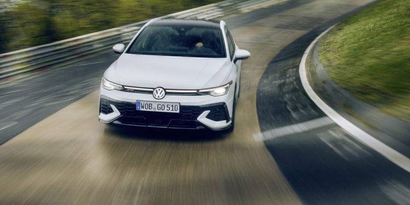 White 2026 Volkswagen Golf GTI with red grille accent driving through a corner on a race track under an overcast sky
