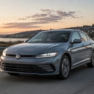 Gray 2026 Volkswagen Jetta sedan driving along a coastal highway at sunset with a hillside town and ocean in the background