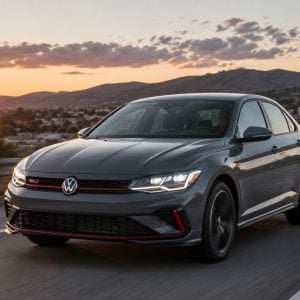 Gray 2026 Volkswagen Jetta GLI sedan with GLI grille badge driving on a highway at sunset with mountains in the background