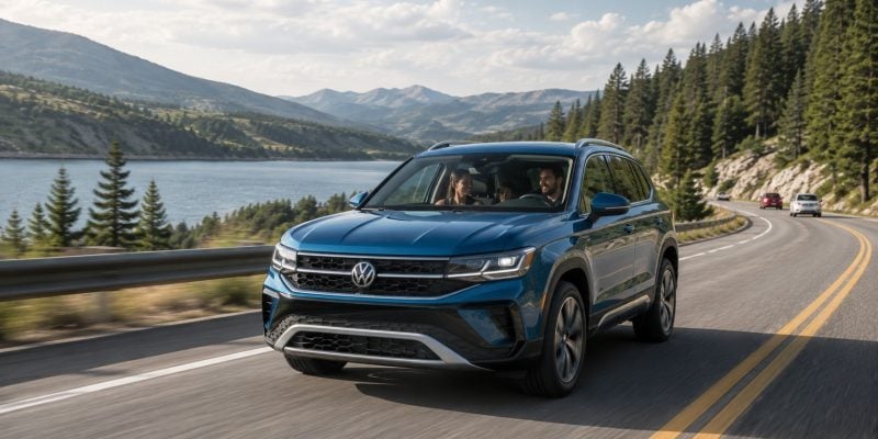 A blue 2026 Volkswagen Taos SUV drives along a mountain lake road with a smiling family visible inside, pine trees and mountains in the background.
